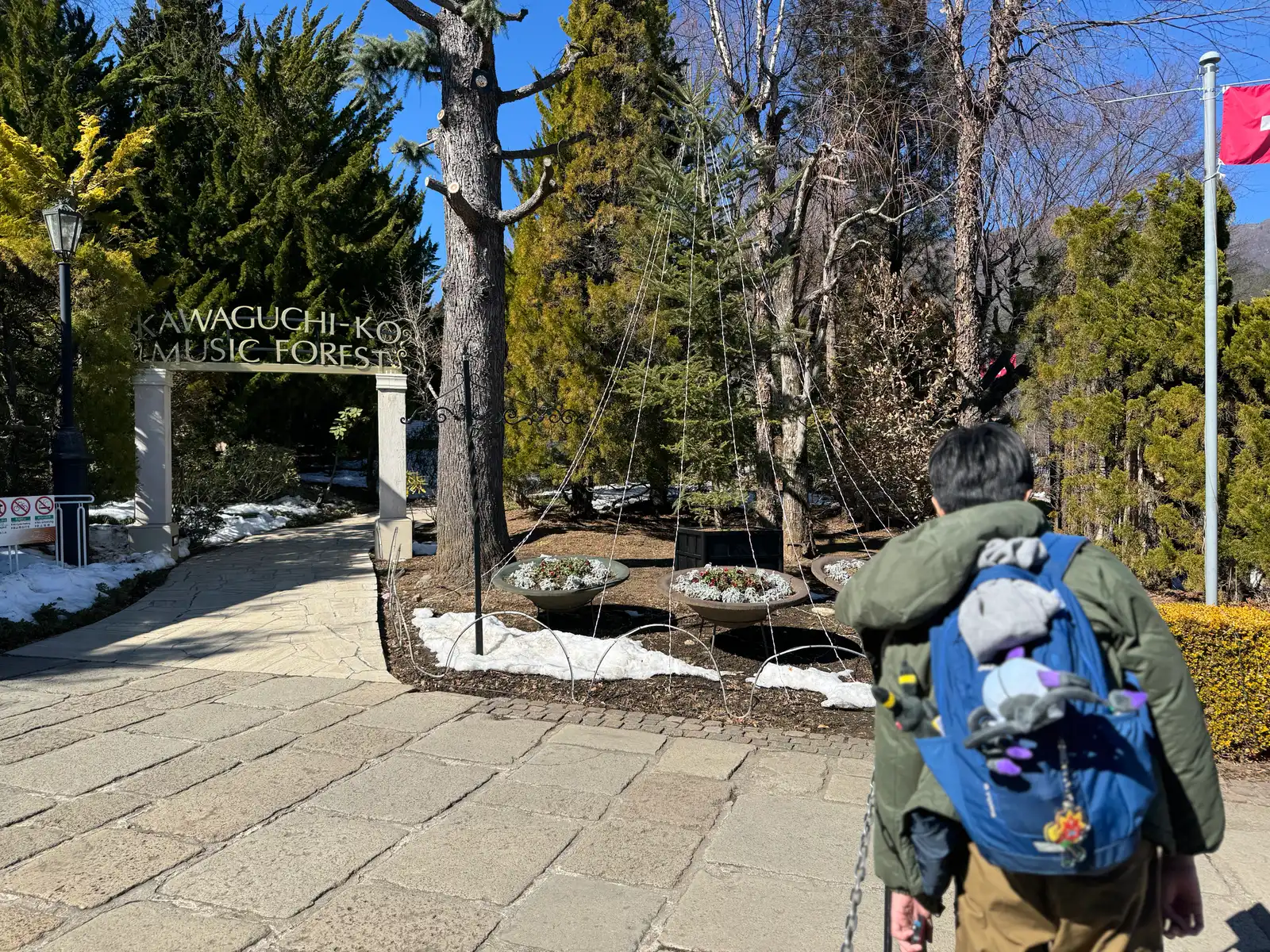 Person in green jacket and blue backpack walks toward Kawaguchiko Music Forest stone sign