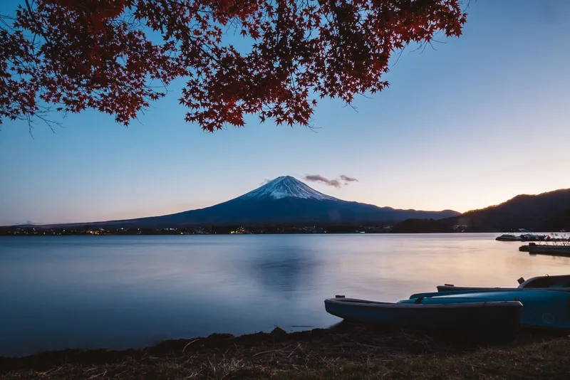 Mt Fuji at dusk across Lake Kawaguchi, red maple leaves above and boats on the shore